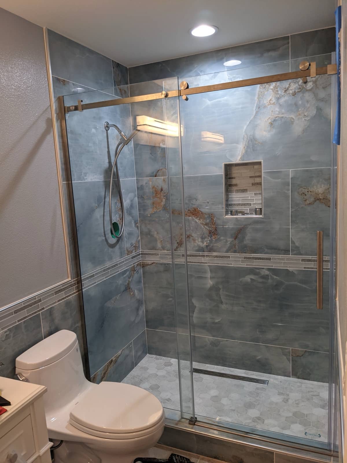 Finished walk-in shower with large-format blue onyx-look porcelain walls, a glass mosaic border and niche, hex mosaic floor, linear drain and frameless sliding glass door